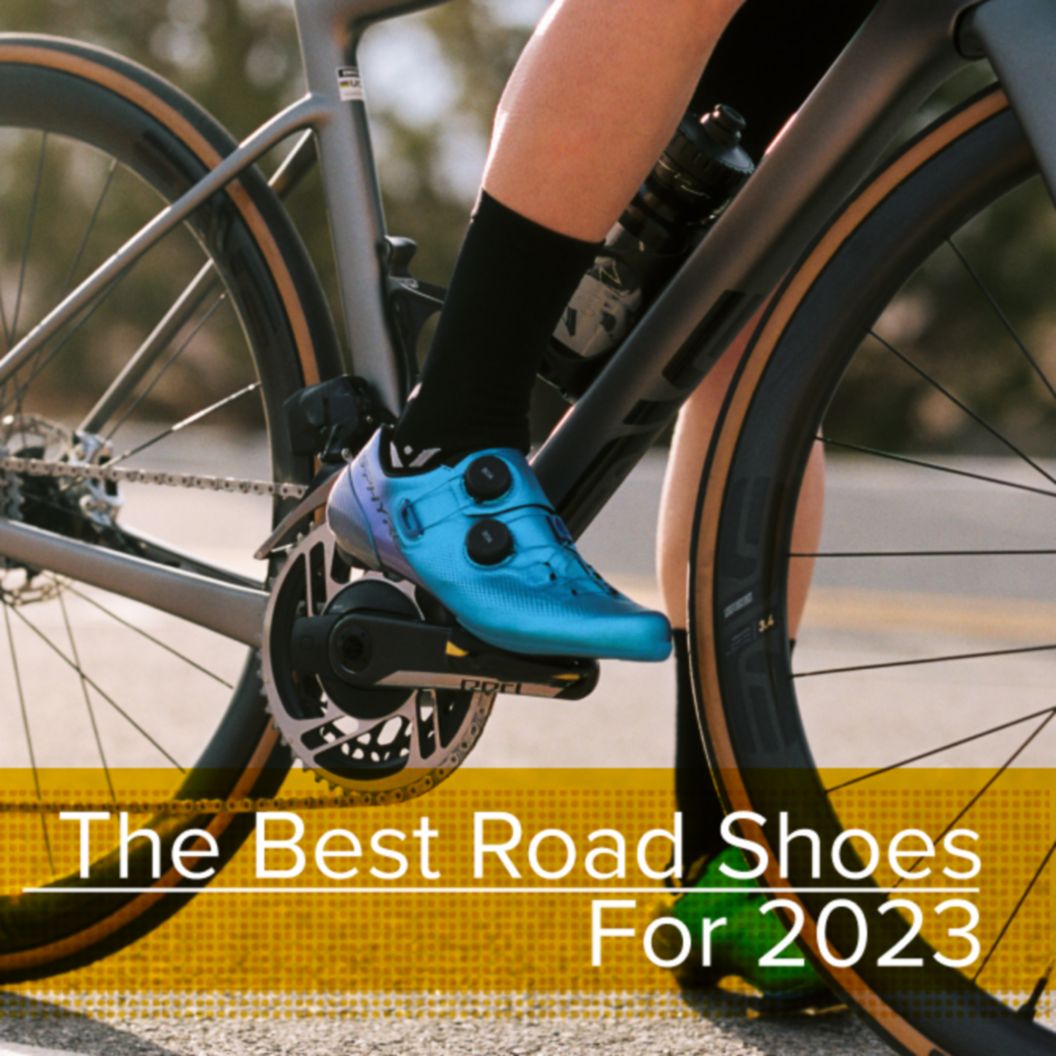 A cyclist in blue Shimano shoes has their right foot clipped into the pedals while standing over their bike. 